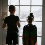 Two small children look through a glass door into the out of focus outside world.