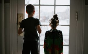 Two small children look through a glass door into the out of focus outside world.