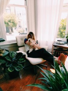 Woman in a beige apartment with many plants, sitting on a chaise lounge and hugging a small white dog. 