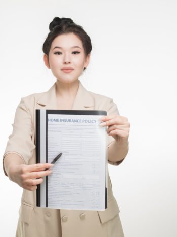 Photograph of a woman in a beige jacket pointing to a home insurance application. She stands in front of a plain white background.